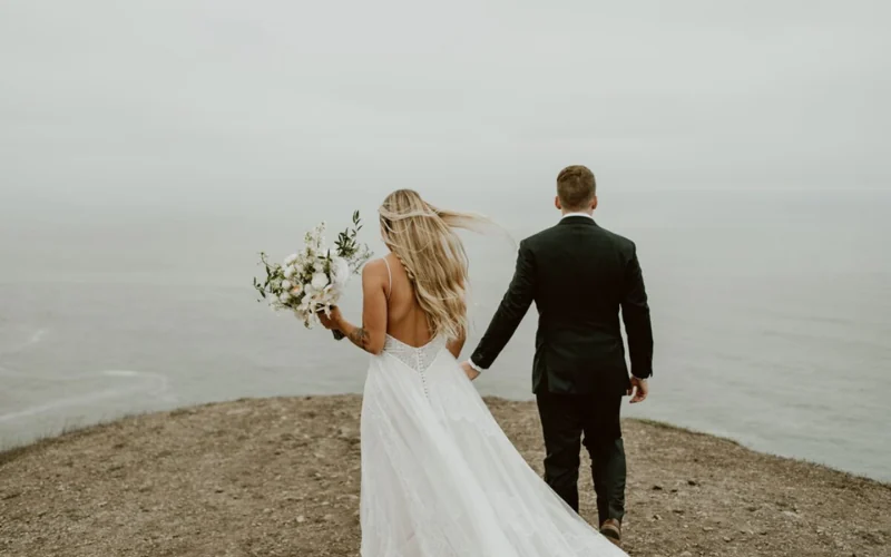 A bride and groom walking towards the sea on a clifftop, her long hair worn loose and lifting in the onshore wind