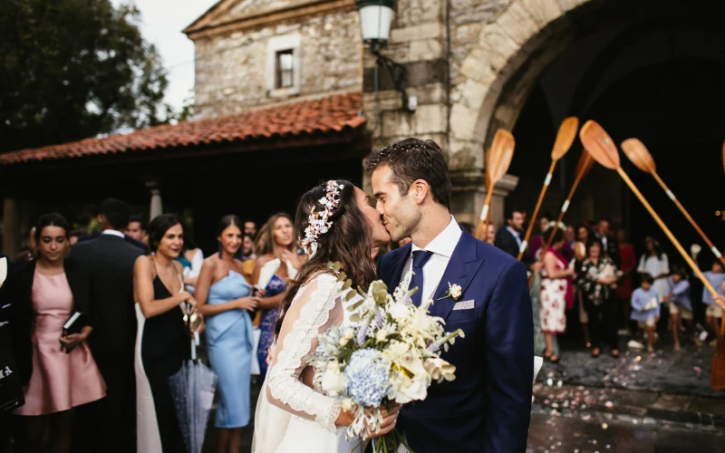 A bride and groom kissing outside a Spanish stone church as their wedding guests look on and confetti covers the steps