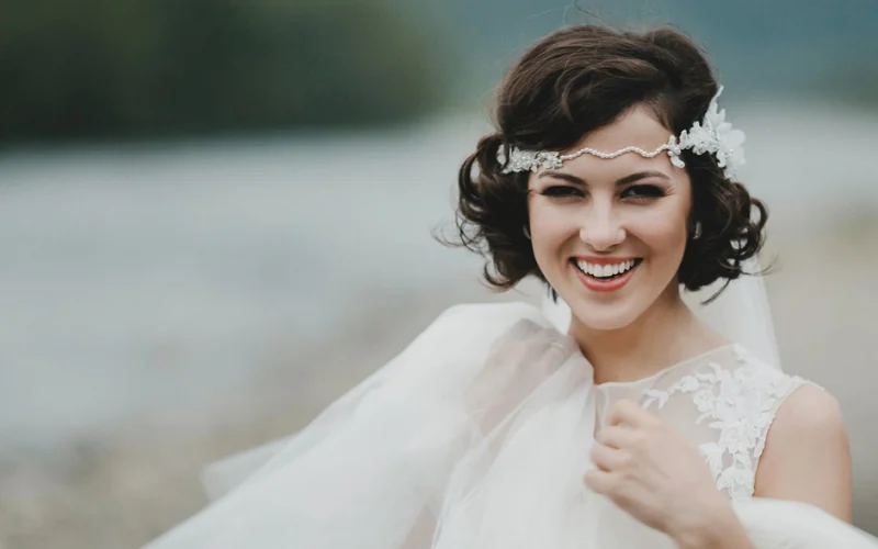 A smiling bride with a short dark curly bob, wearing a jewelled browband headpiece and a soft tulle veil