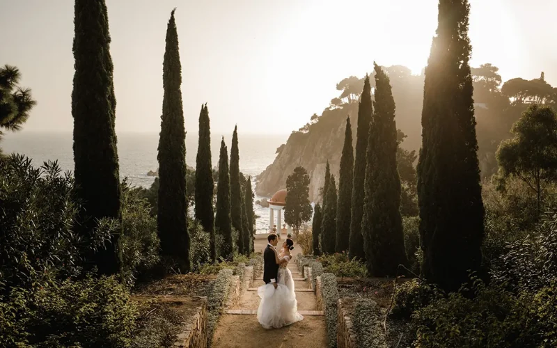 A bride and groom on a cypress lined path leading down towards the sea at a coastal wedding venue in Spain at sunset