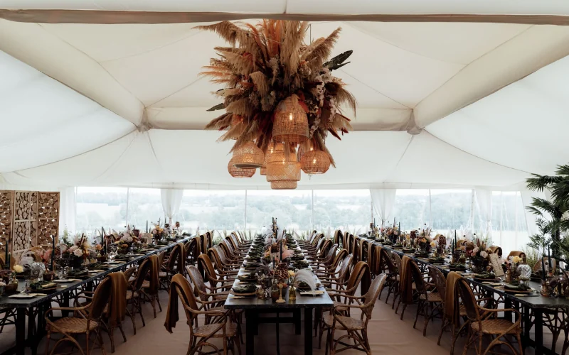 A dried pampas and rattan chandelier suspended from the centre of a marquee ceiling above long wedding tables