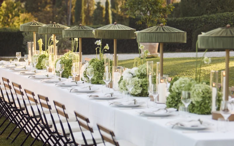 Green pleated lampshades on tall brass stands running down a long white wedding table set with white hydrangeas and candles