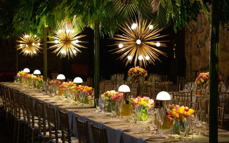 Gold starburst light fittings hanging above a long wedding table at night, with small dome lamps and bright seasonal flowers