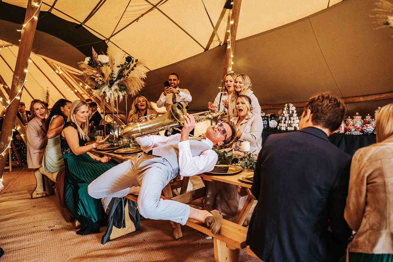 A wedding saxophonist leaning back across a bench mid performance while seated guests laugh and film him at a reception